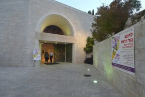 תצלום של חזית מרכז מורשת מנחם בגין בירושלים בשעת ערב, כשעל קיר האבן הימני תלויה כרזה גדולה של אירוע השקת הספר "לרכוב נגד הרוח" ובחזית הכניסה מוצב שלט מכוון קטן עם אותה הכרזה