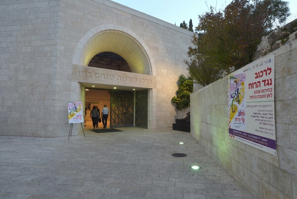 תצלום של חזית מרכז מורשת מנחם בגין בירושלים בשעת ערב, כשעל קיר האבן הימני תלויה כרזה גדולה של אירוע השקת הספר "לרכוב נגד הרוח" ובחזית הכניסה מוצב שלט מכוון קטן עם אותה הכרזה
